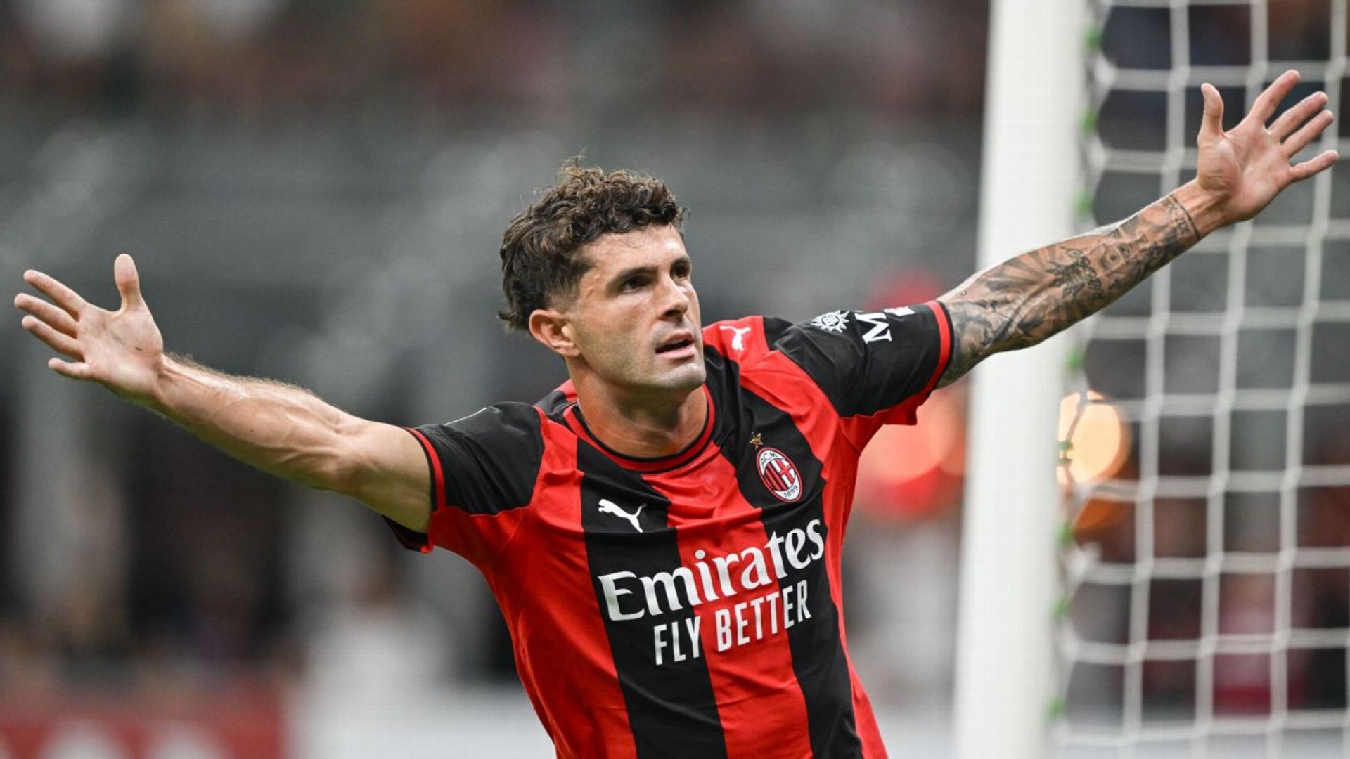 Christian Pulisic in AC Milan's red-and-black home kit, arms outstretched by the goal at San Siro