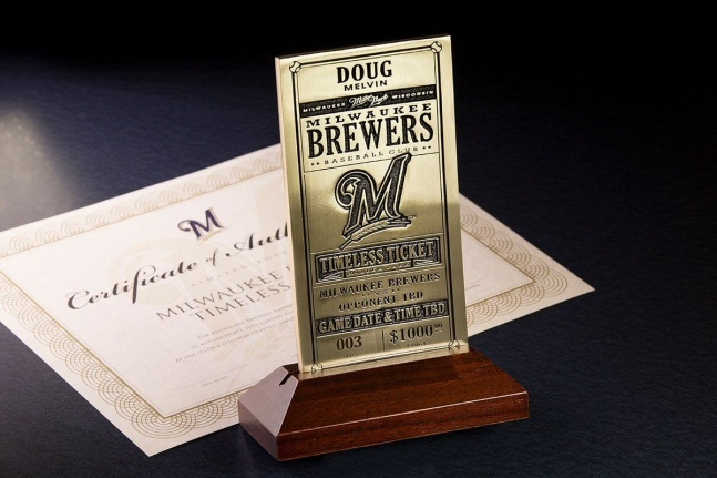 The Milwaukee Brewers' brass Timeless Ticket on its wooden stand — engraved with a holder's name, numbered 003 and priced at $1,000, 'Game Date & Time TBD' — beside the program's certificate, in the club's promotional photograph from the 2015 sale