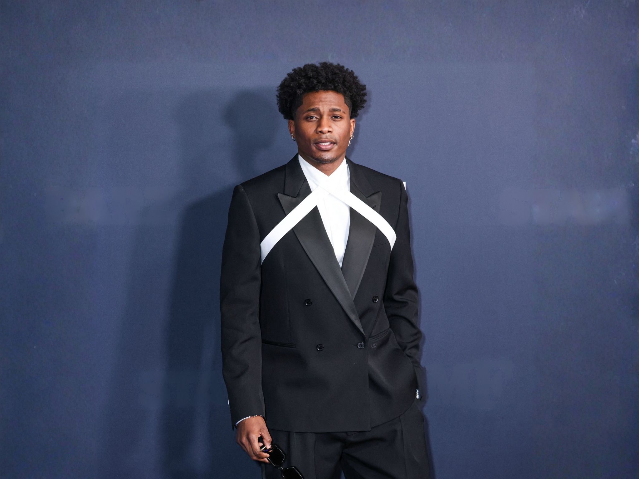 Da'Vinchi in a black double-breasted suit with a white cross-strap shirt, photographed against a dark blue backdrop — red-carpet file photo