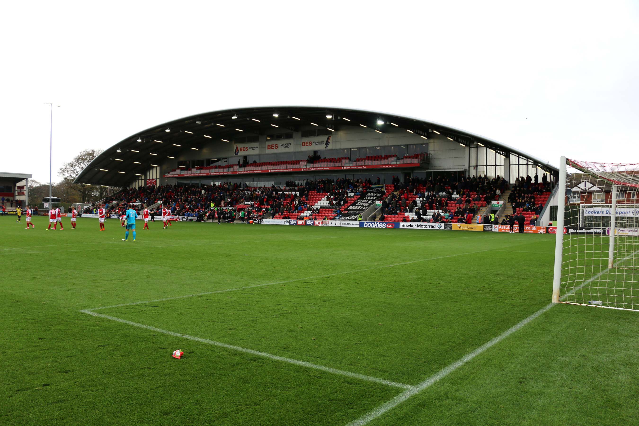 The Parkside Stand at Highbury Stadium, Fleetwood, during a Fleetwood Town match — the home crowd in the red seats, Fleetwood players in red and white on the pitch, and a goal in the foreground — 2017 file photo