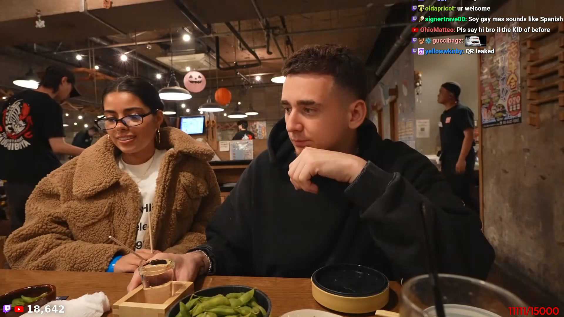 Lacy seated at the restaurant table in Japan during his October 28, 2026 livestream, chin on fist mid-broadcast, dishes on the table in front of him, restaurant interior behind, and the stream's live chat visible at top-right including a viewer's 'QR leaked' message