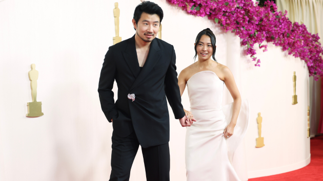 Simu Liu and Allison Hsu walk the red carpet hand in hand at the 2024 Academy Awards, him in black and her in a white one-shoulder gown.