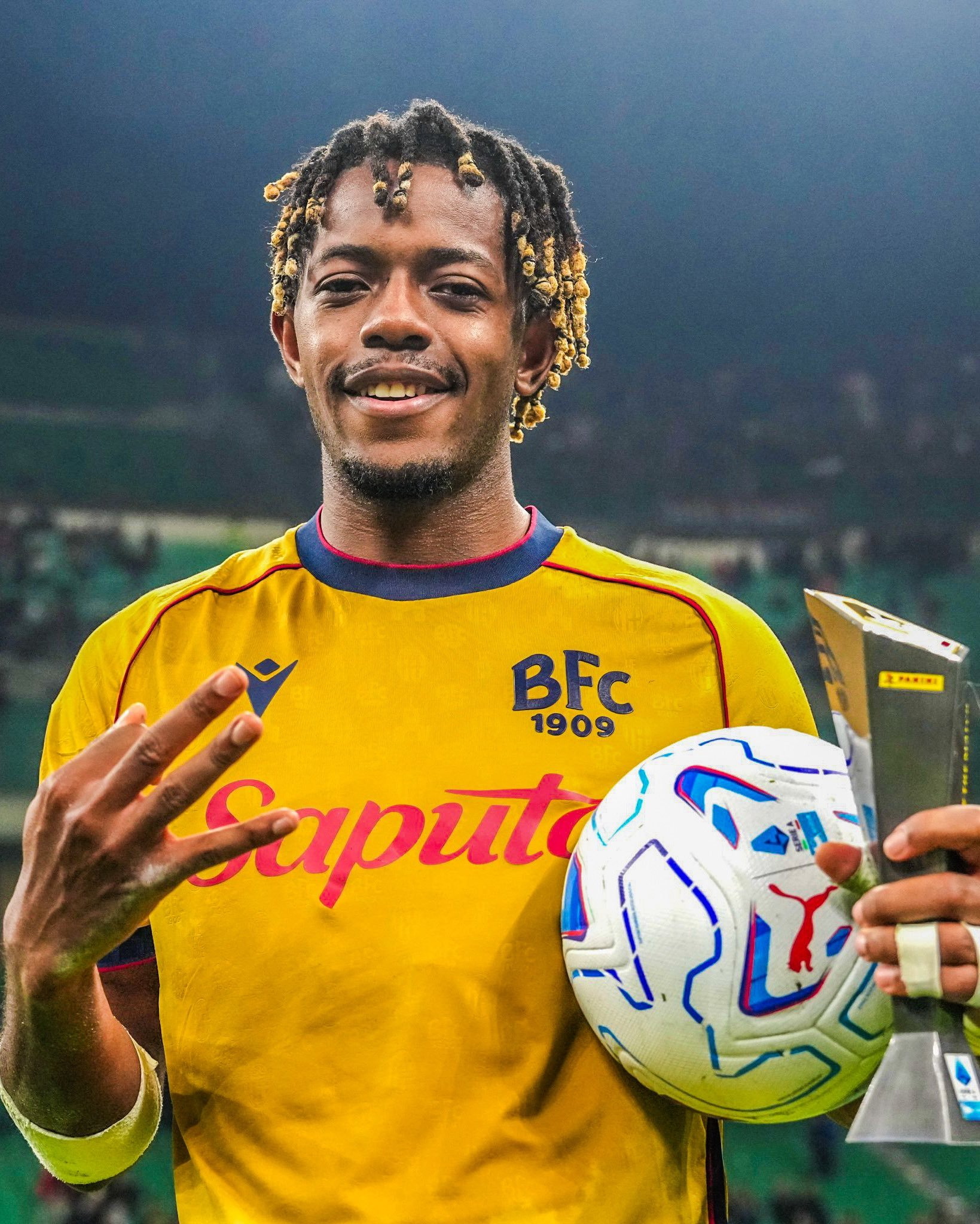 Samuel Mbangula of Bologna, in the yellow away shirt, holding the Serie A match ball and the Panini Player of the Match award and holding up three fingers after his hat trick against AC Milan at San Siro, October 28, 2026