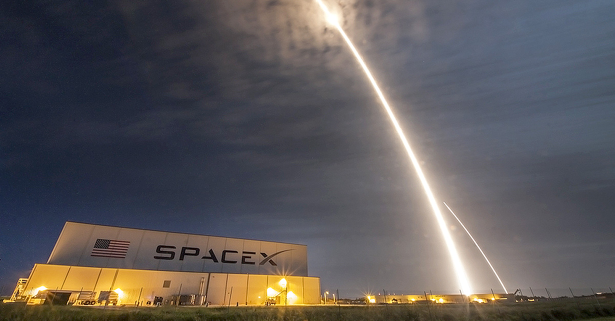 A SpaceX rocket’s bright launch trail arcing over a SpaceX facility at night