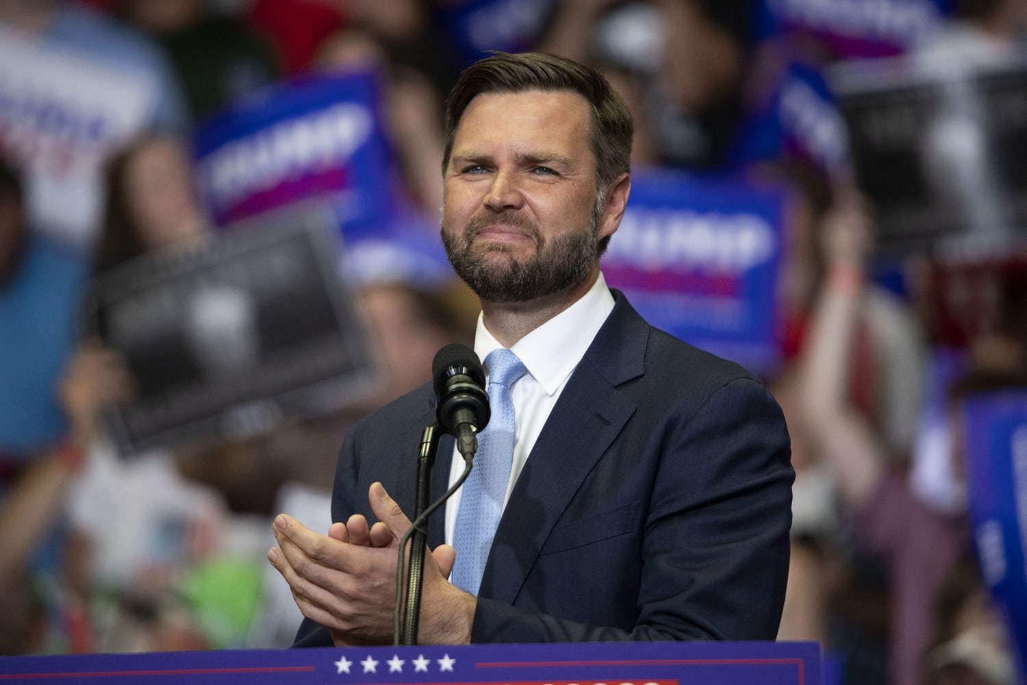 Vice President JD Vance speaking at a campaign podium with supporters and signs behind him.
