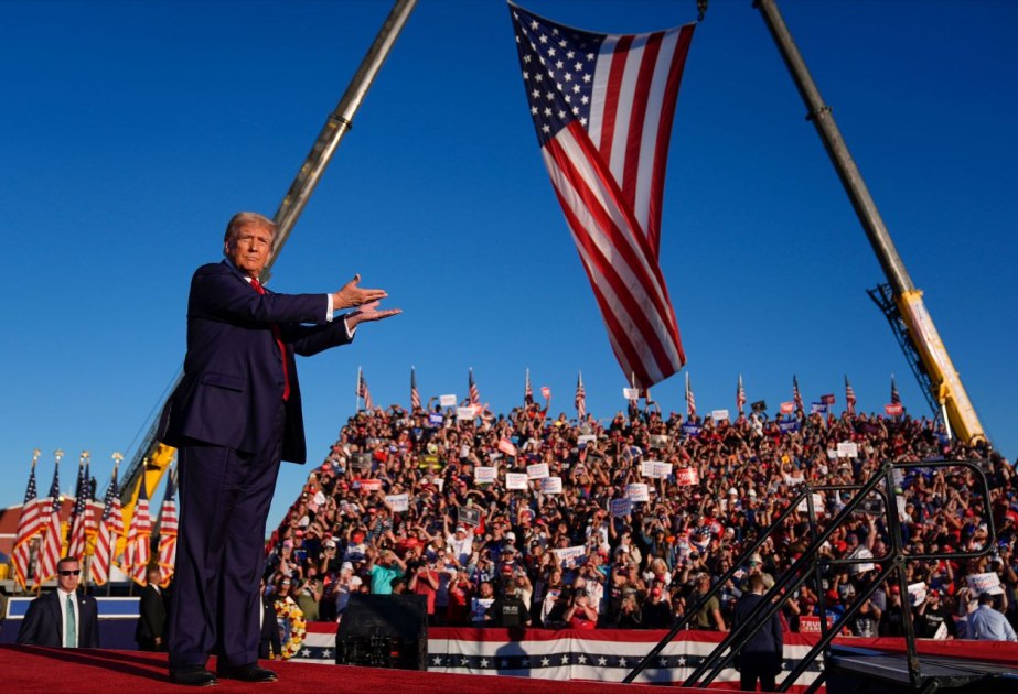A packed campaign rally crowd with American flags under a blue sky