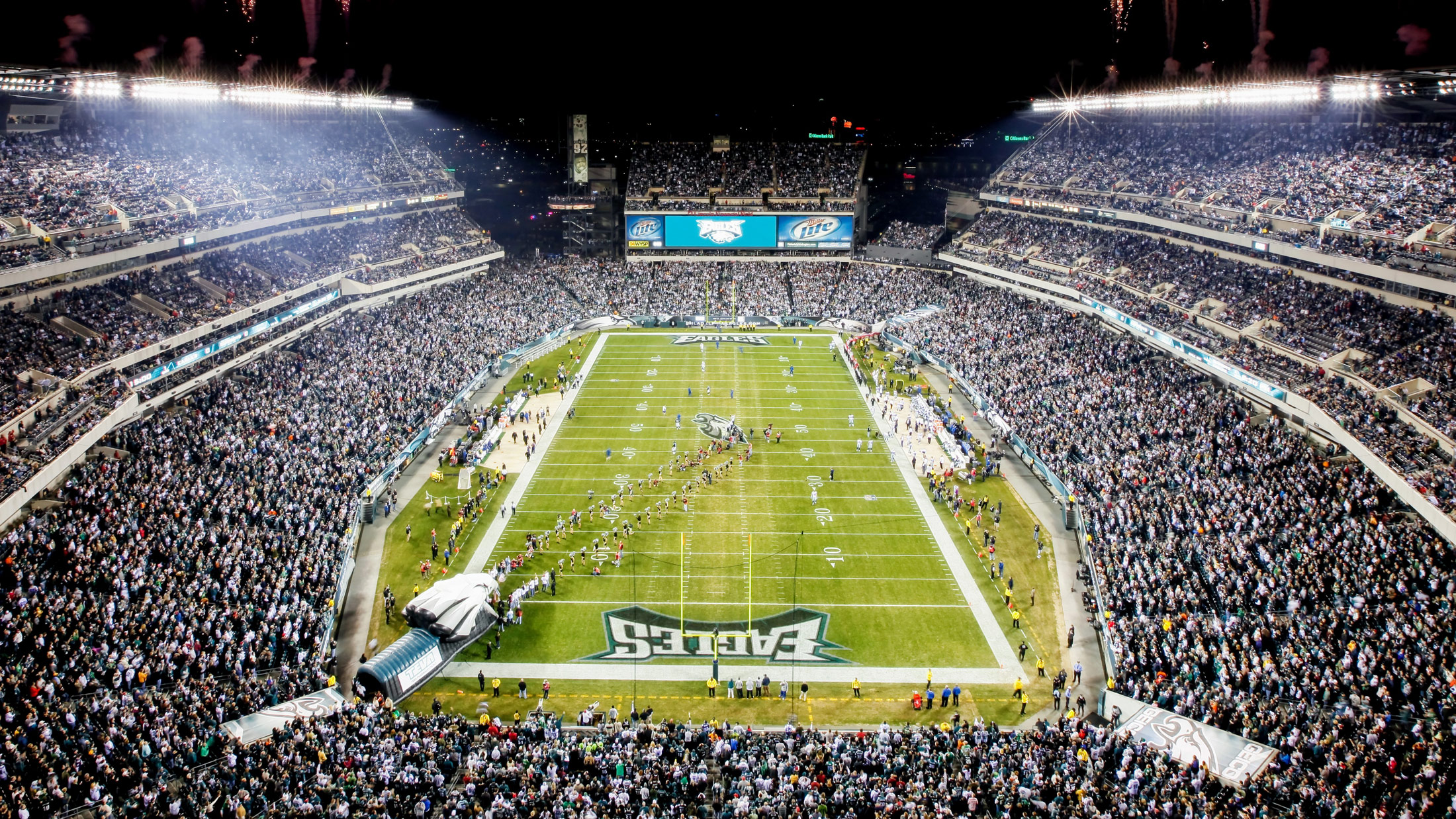 Aerial night view of Lincoln Financial Field in Philadelphia, packed with fans under the stadium lights with fireworks overhead.