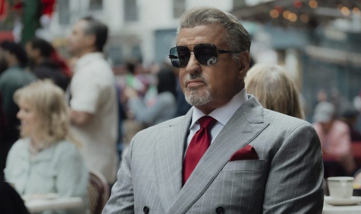 Sylvester Stallone as Dwight Manfredi in Tulsa King, wearing sunglasses and a gray pinstripe suit with a red tie