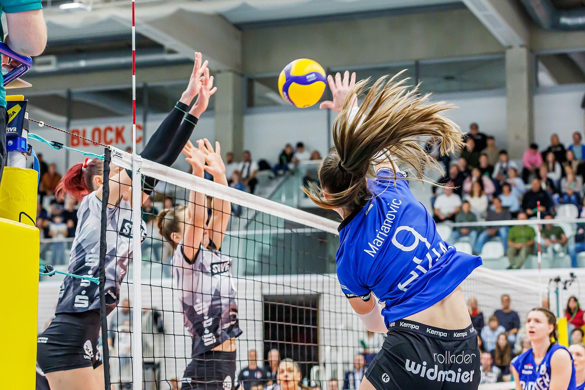 A women's volleyball player mid-attack against a double block in a packed indoor arena