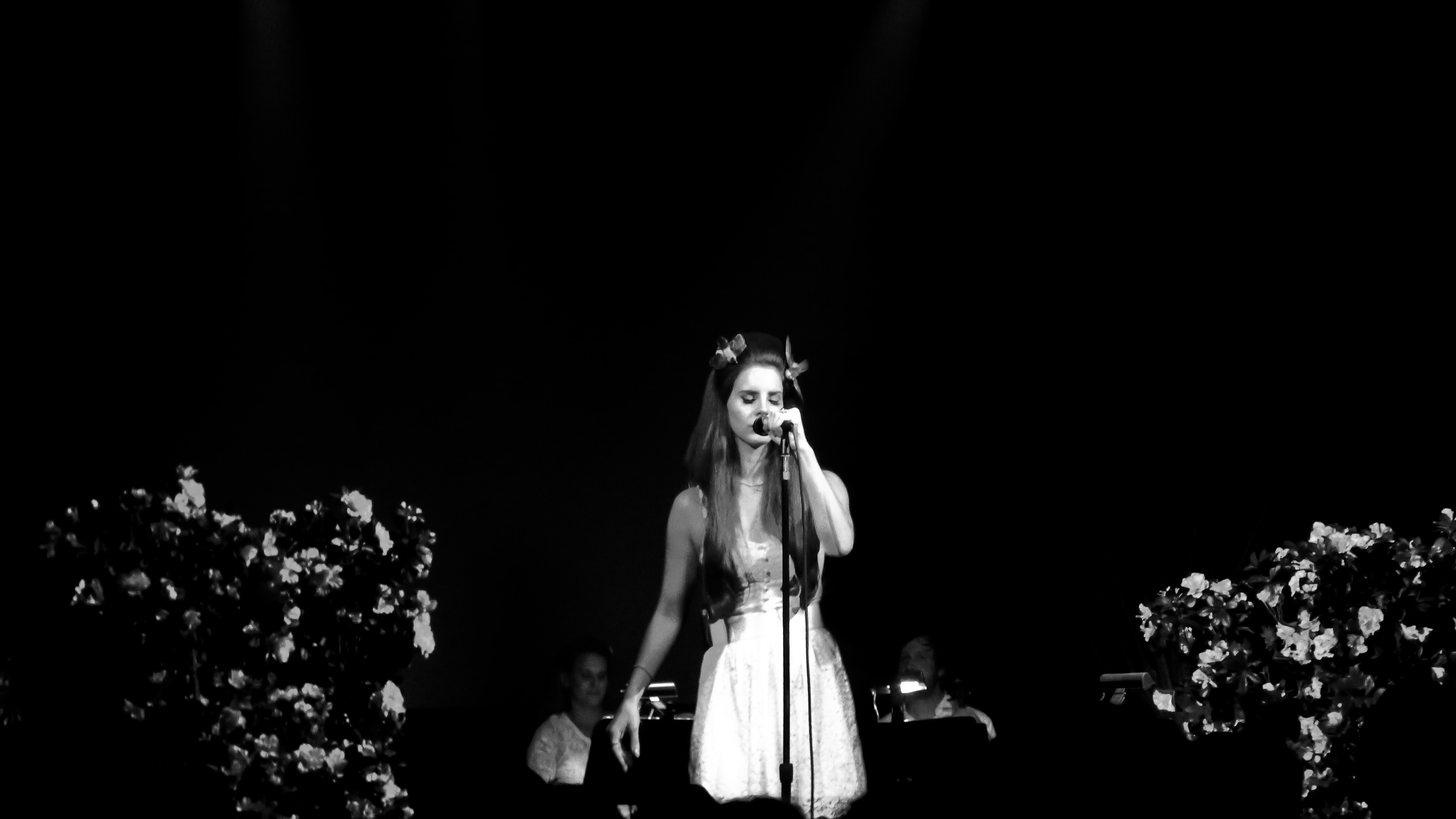 Lana Del Rey performing live, in a white dress at a microphone stand between floral arrangements, photographed in black and white in New York